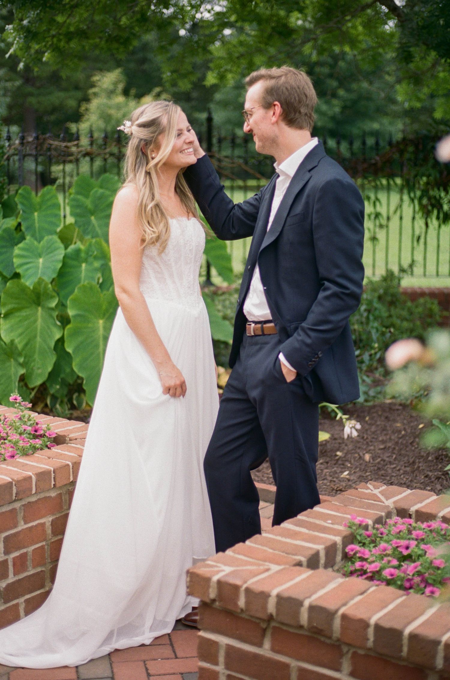 Bride in white gown and groom in navy suit share a tender moment in a lush garden setting.