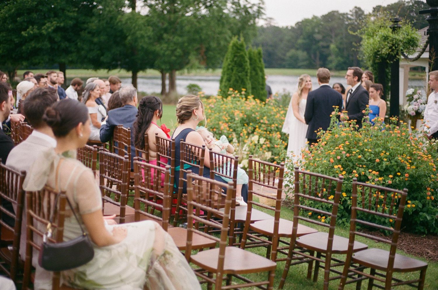 Outdoor wedding ceremony with guests seated in wooden chairs beside colorful garden flowers near a scenic lake.