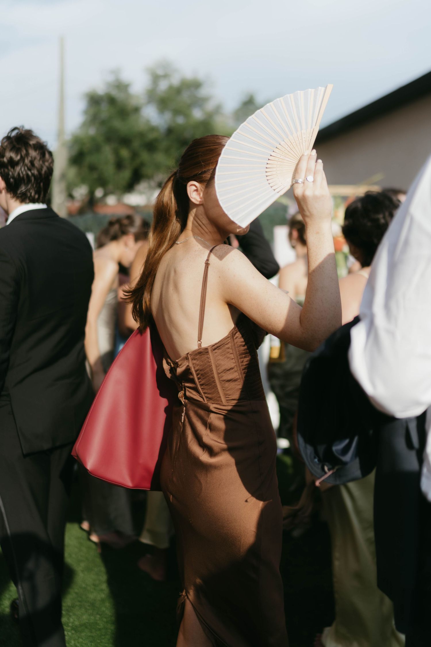 A wedding guest in a brown dress holds up a white folding fan during an outdoor reception.