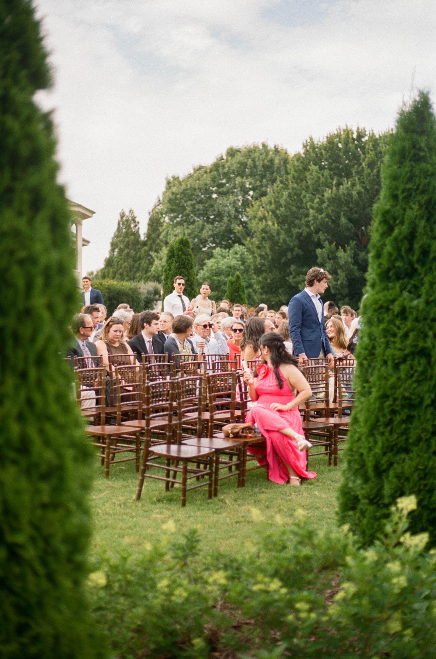 Guests seated in wooden chairs at an elegant outdoor wedding ceremony surrounded by lush green hedges and trees.