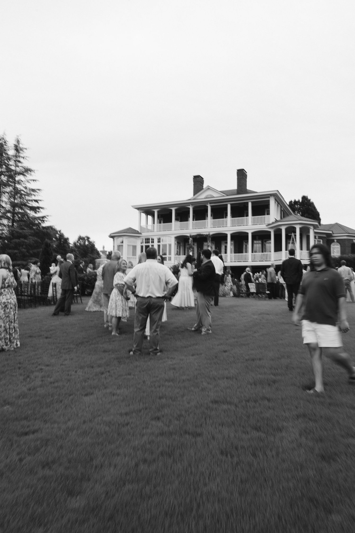 Wedding guests gather on lawn before grand white two-story plantation-style manor at dusk.