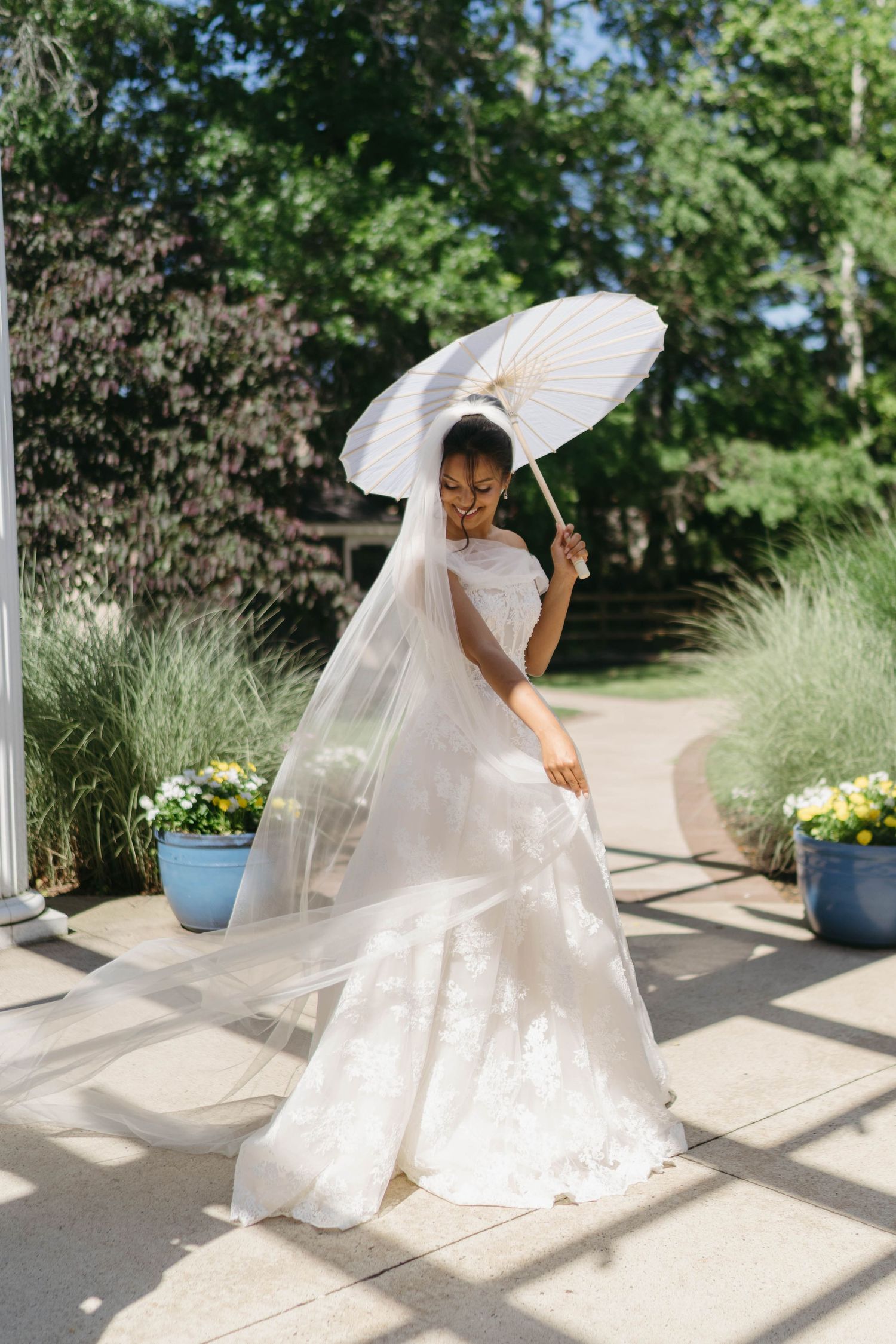 Bride in white lace gown holding a white parasol umbrella outdoors on a sunny day with lush green trees.