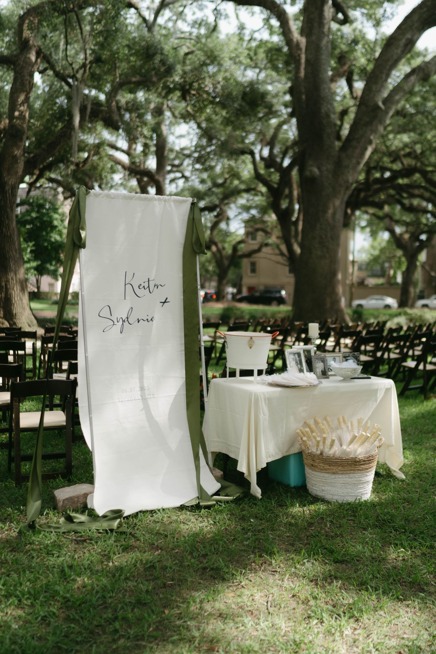 White chairs arranged for an outdoor wedding ceremony under large oak trees.