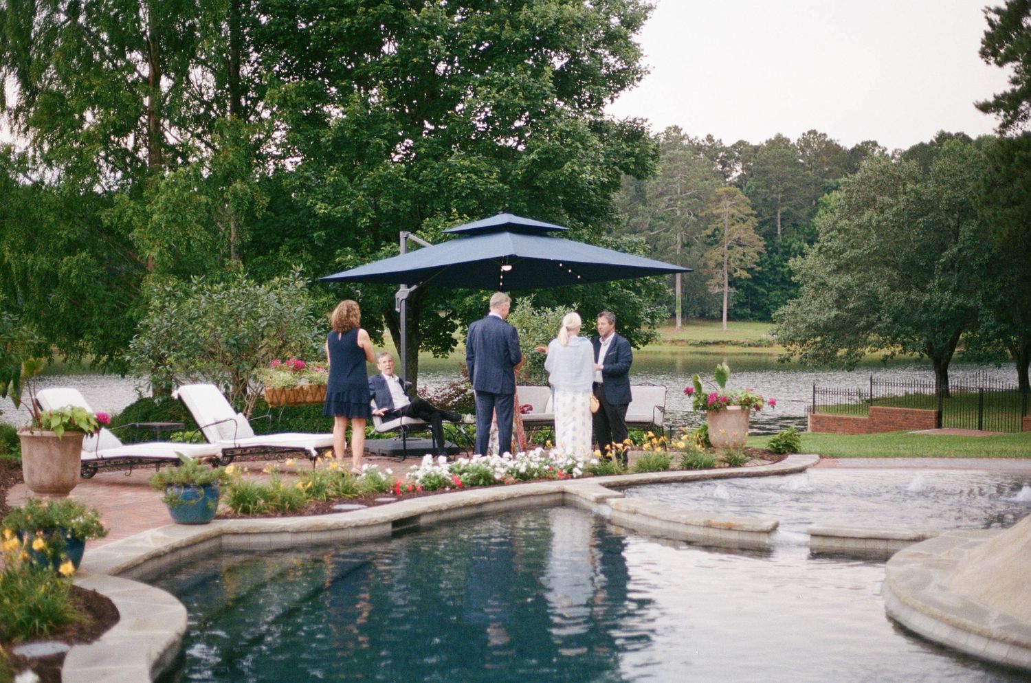 Elegant outdoor poolside gathering with guests under a navy umbrella gazebo surrounded by lush gardens and a serene lake backdrop.