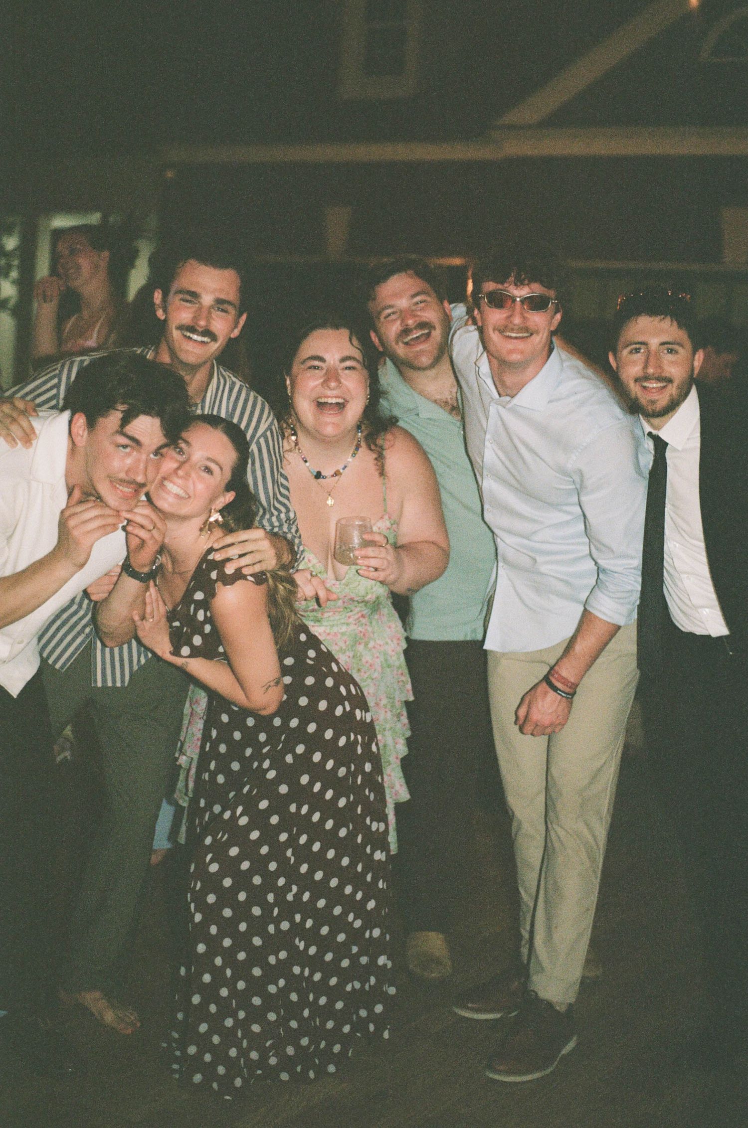 Group of joyful wedding guests laugh and pose together during a lively outdoor evening wedding reception.
