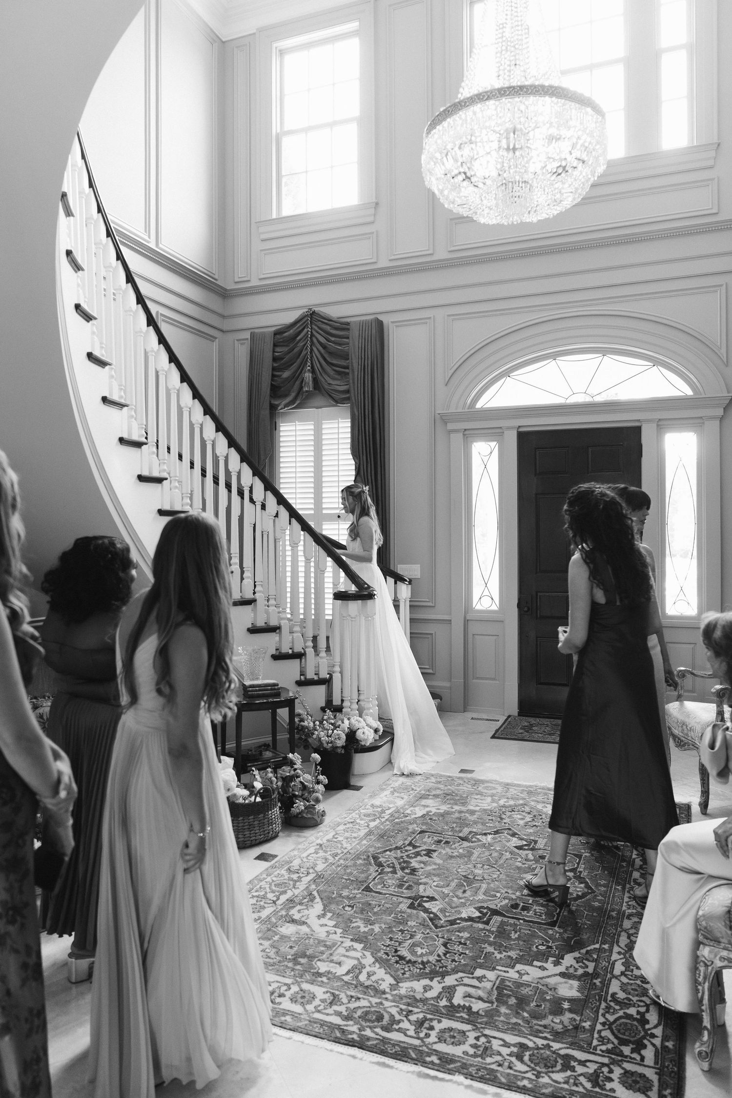 Black and white photo of bride descending grand staircase with bridesmaids in elegant foyer.