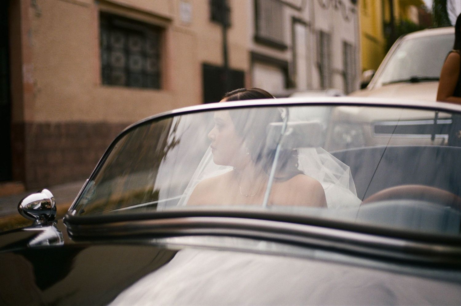 Bride in white dress being assisted into a classic black car.