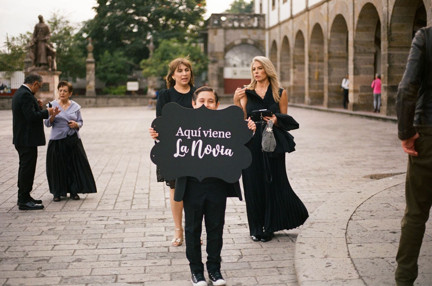 Wedding guests gather on a stone patio outside a historic building with arched architecture.