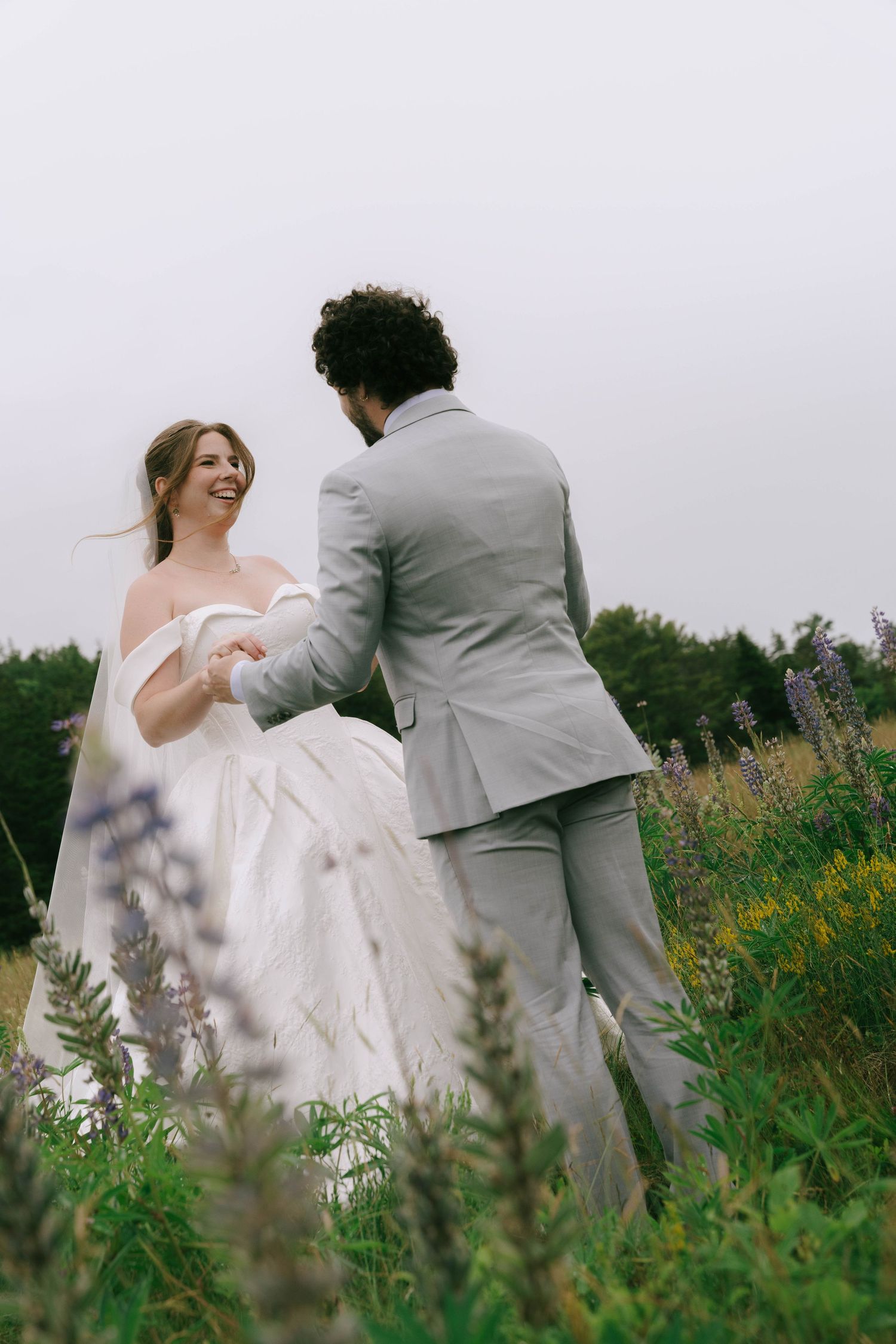A couple in wedding attire shares a playful moment among wildflowers in a field.