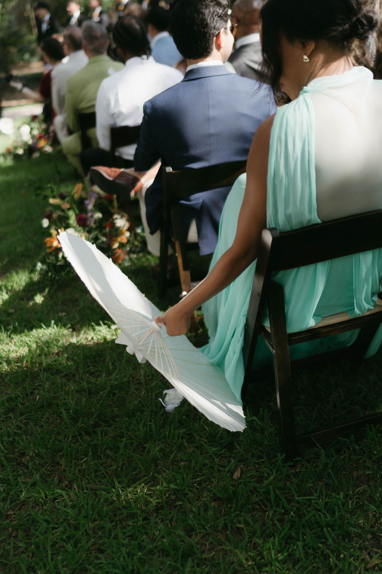 Wedding guests in mint green dresses seated on wooden chairs outdoors.
