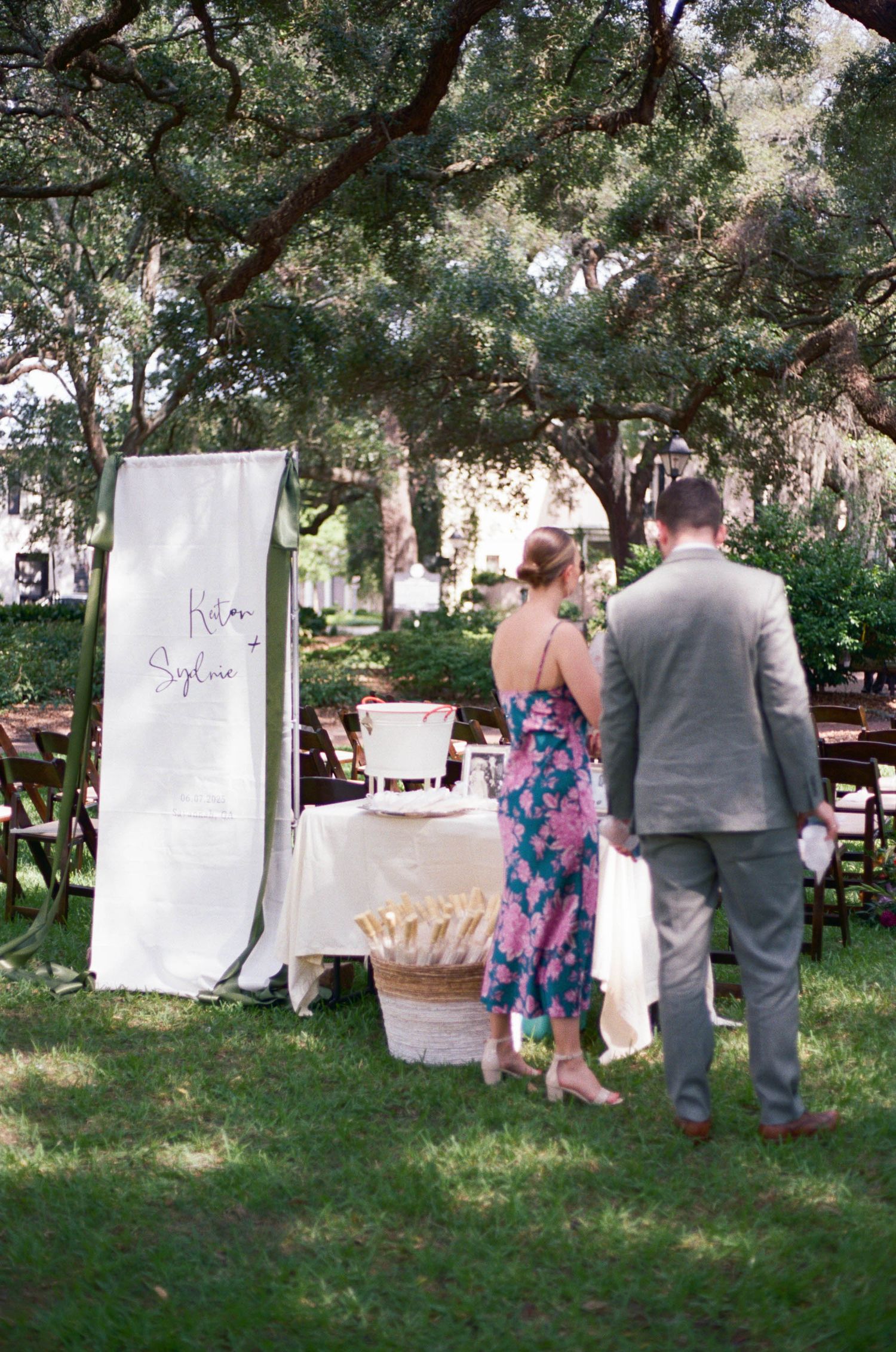 Wedding guests walking past white fabric panels hanging between trees in a garden setting.