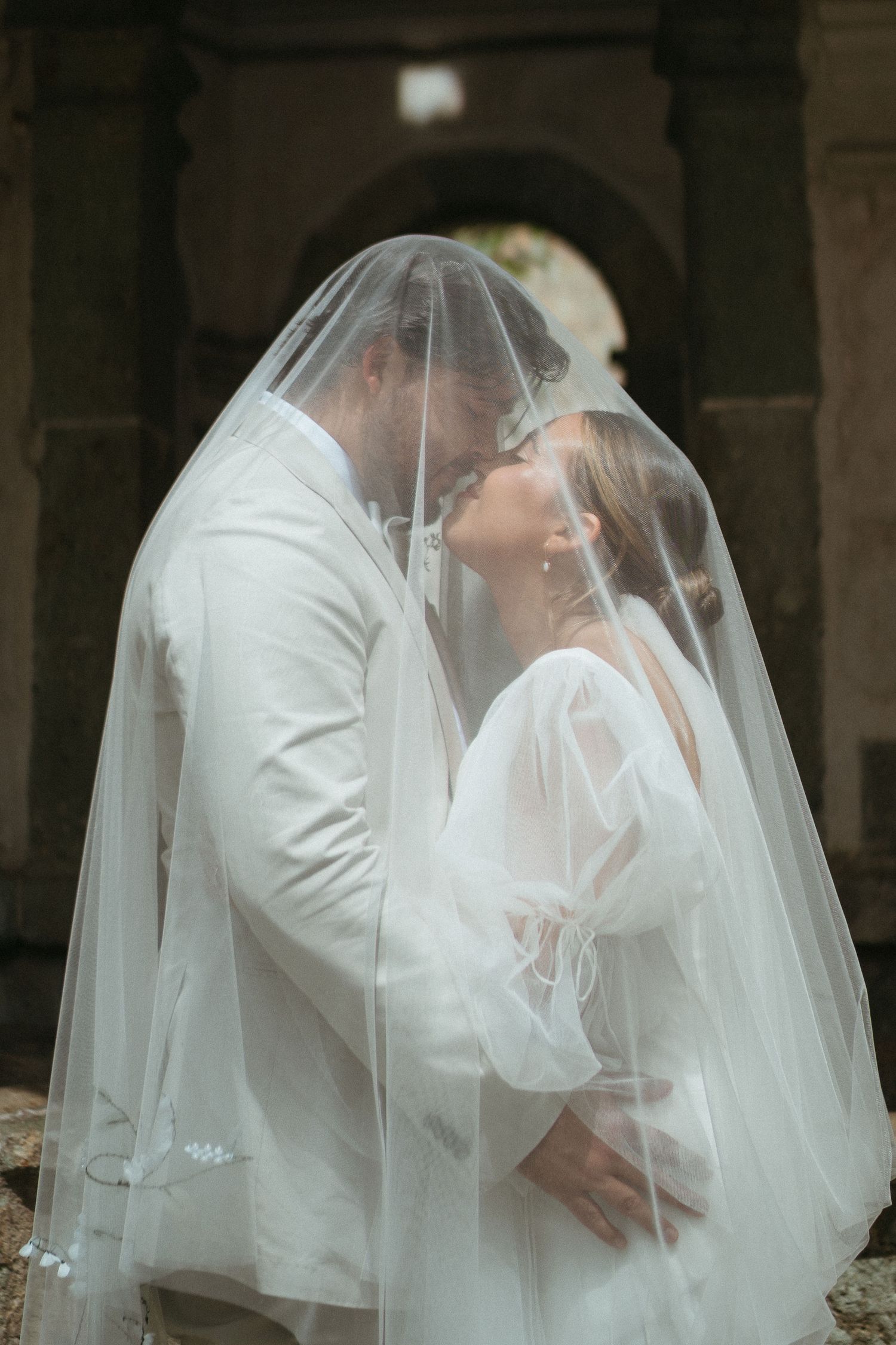 An intimate wedding portrait under stone arches with shared veil and white attire.
