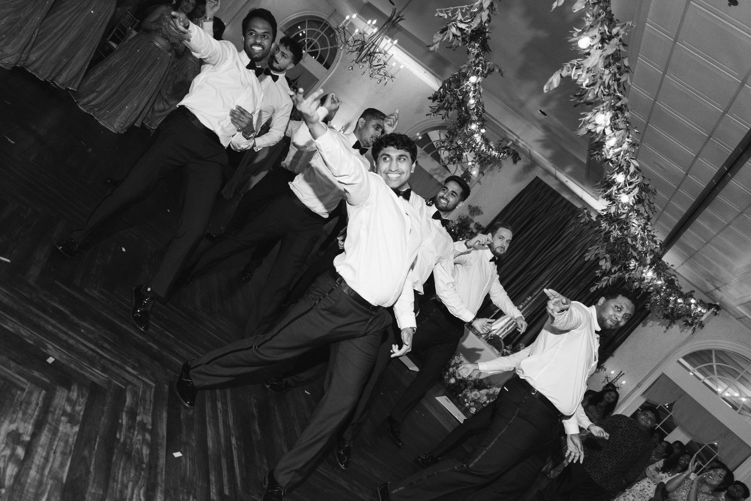Black and white photo of groomsmen dancing and celebrating at a wedding reception in a decorated venue.