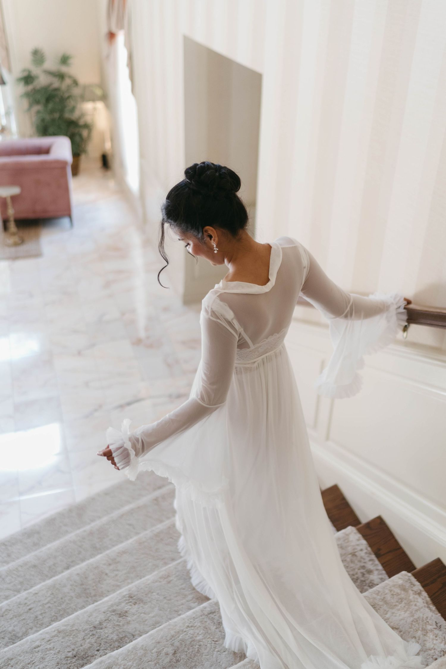 Bride in elegant white long-sleeve chiffon wedding gown descending marble staircase, hair in updo, viewed from behind.