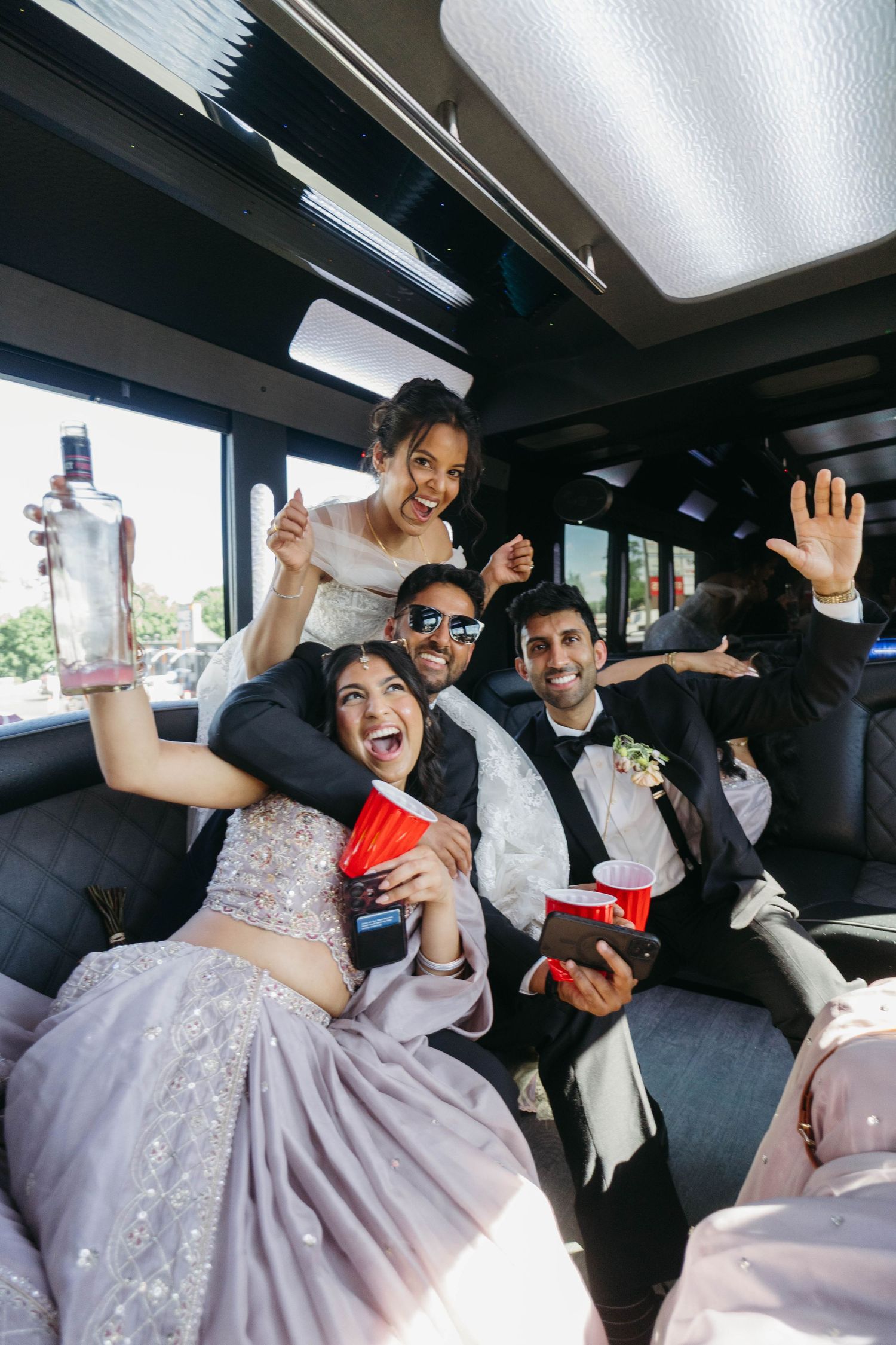 Wedding party celebrating in a party bus, holding red cups and a vodka bottle, dressed in formal attire.