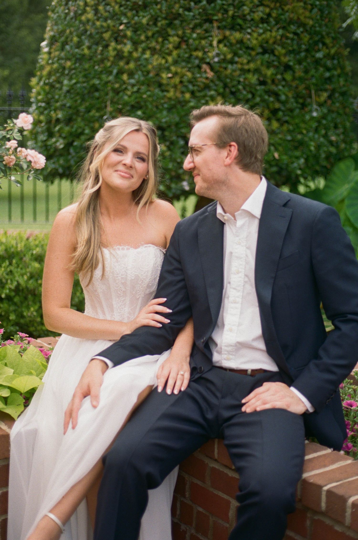 Bride in strapless lace gown sits with groom in navy suit on brick wall surrounded by lush garden greenery.