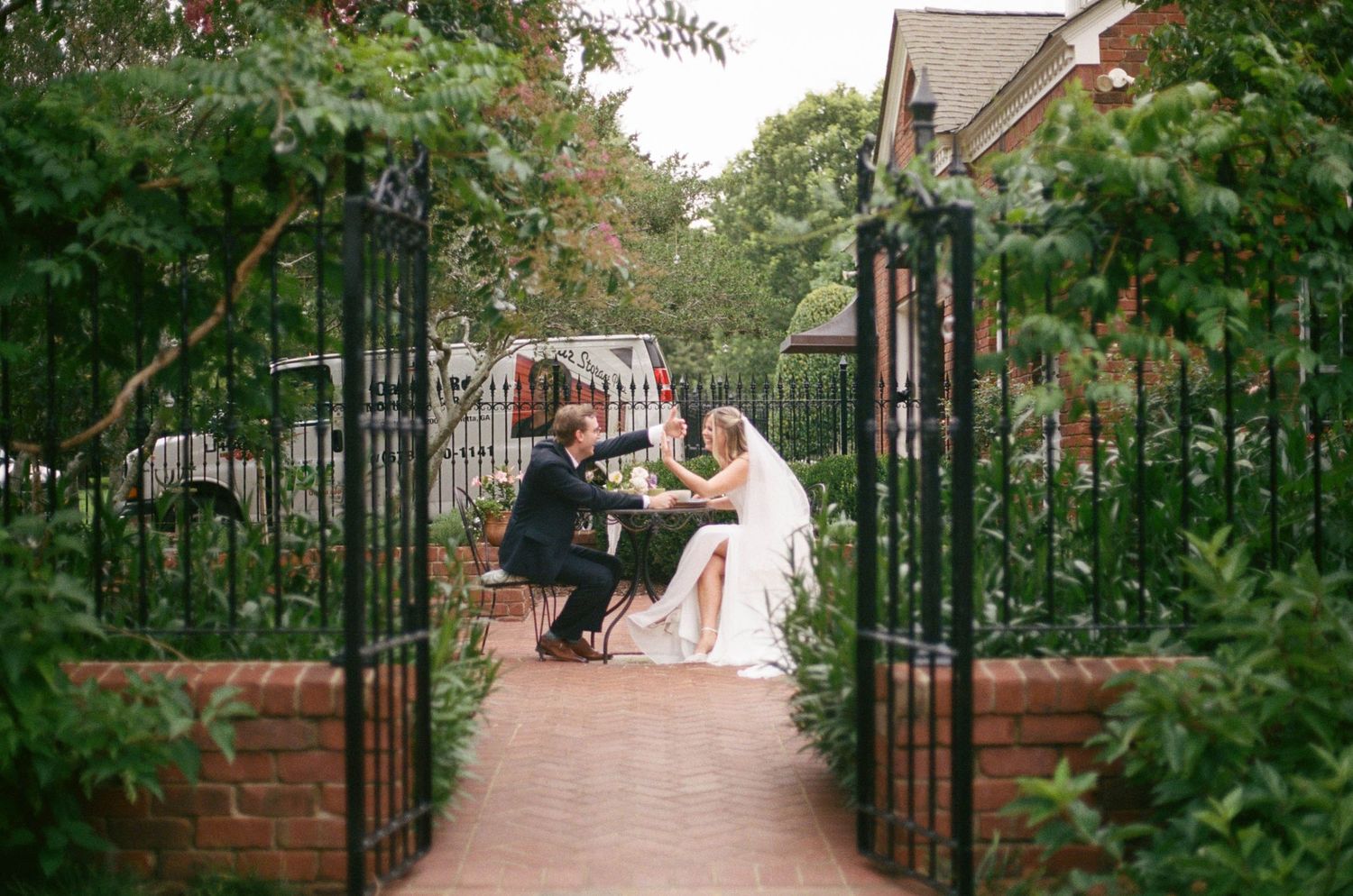 Bride and groom seated at a table in a lush garden courtyard framed by iron gates on their wedding day.