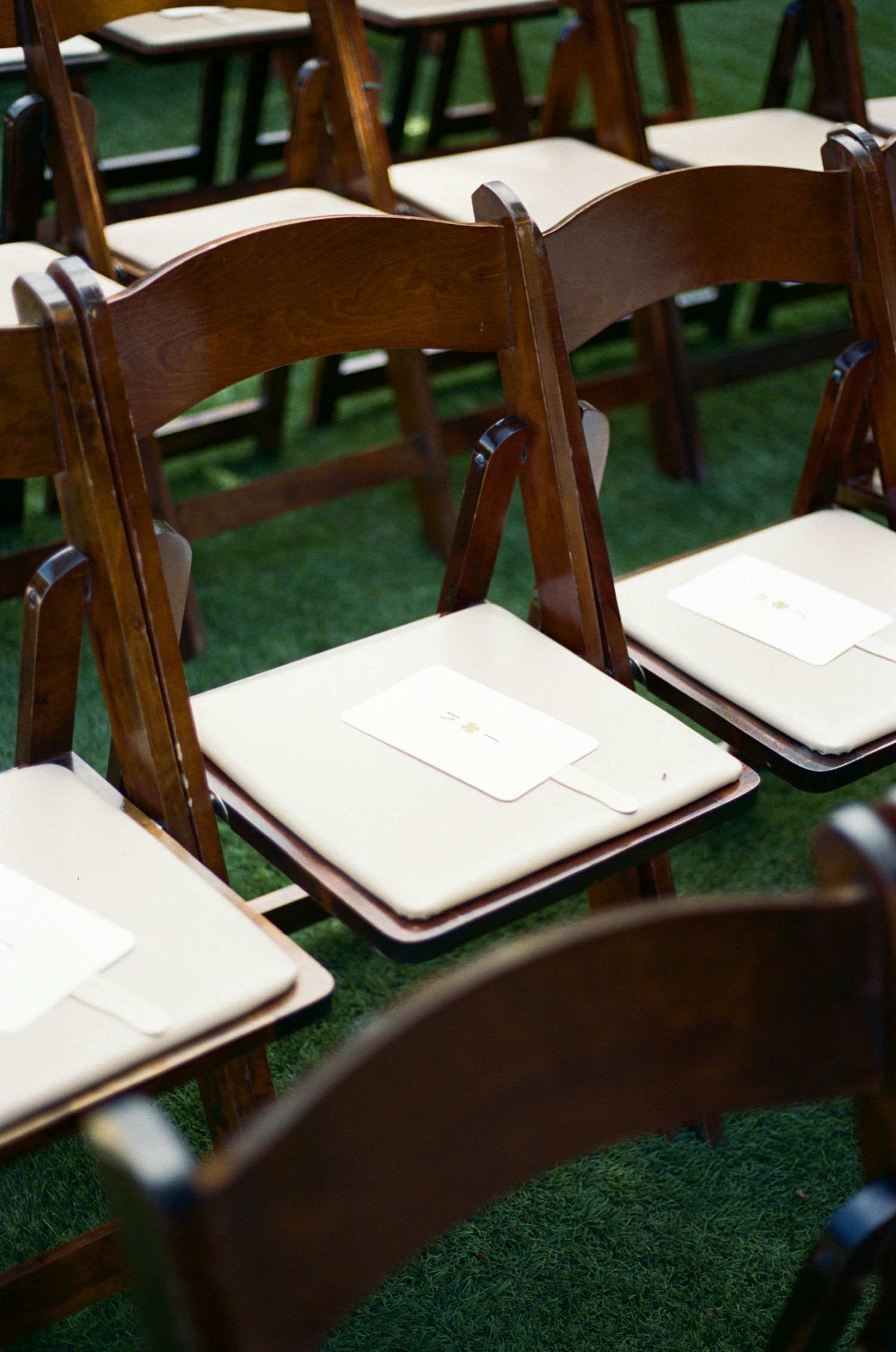 Brown wooden folding chairs with white cushions arranged on green carpet for an outdoor ceremony.