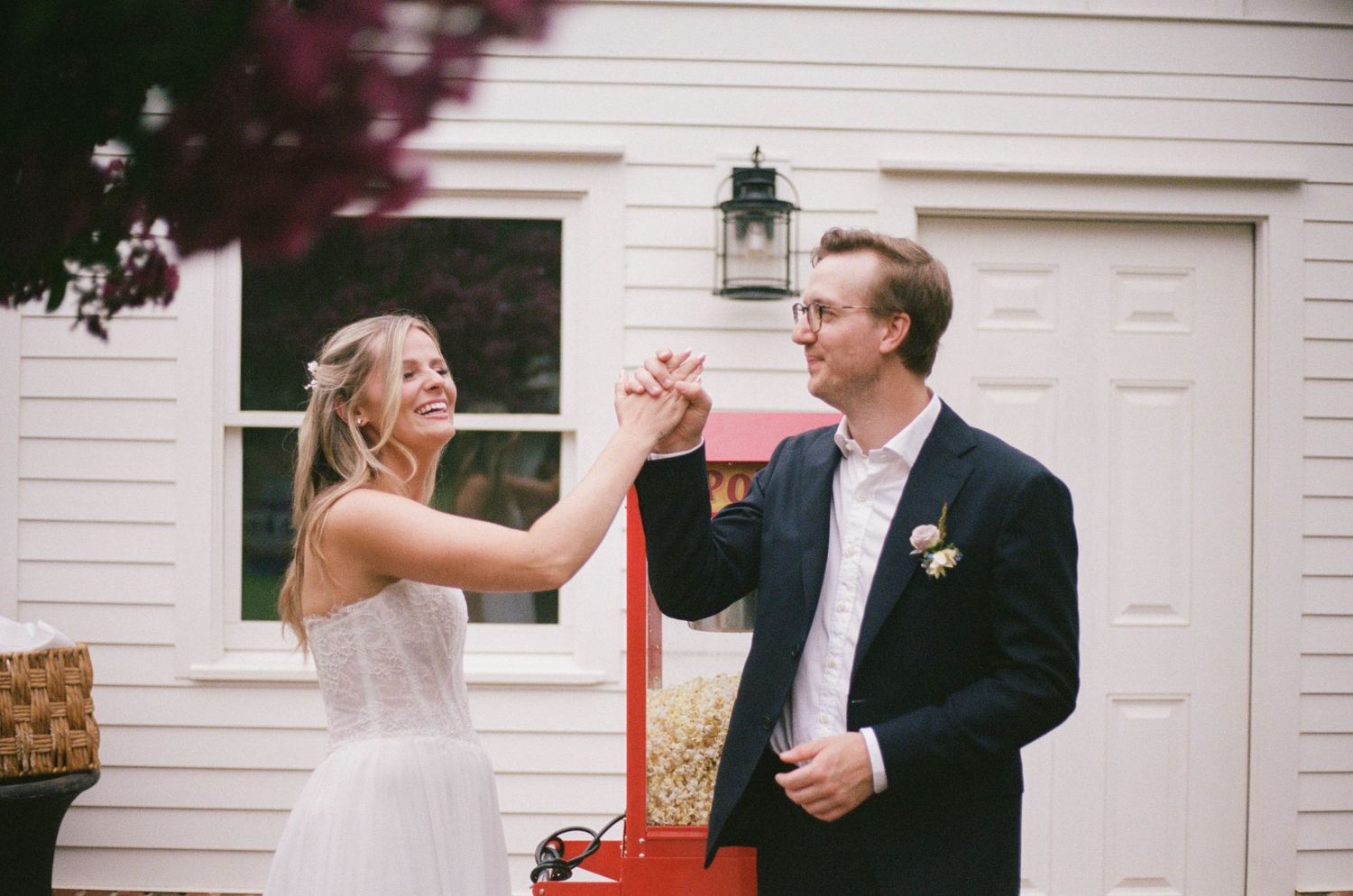 Bride and groom smiling and holding hands outdoors near a white building during their wedding celebration.