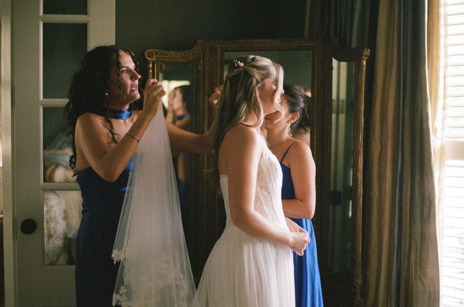 Mother and bridesmaid help bride adjust her veil and hair before the wedding ceremony indoors.
