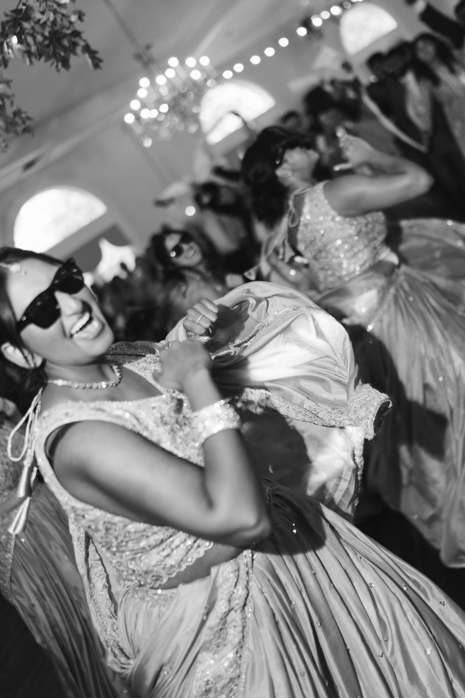 Black and white photo of guests in Indian attire dancing joyfully at wedding reception under string lights.