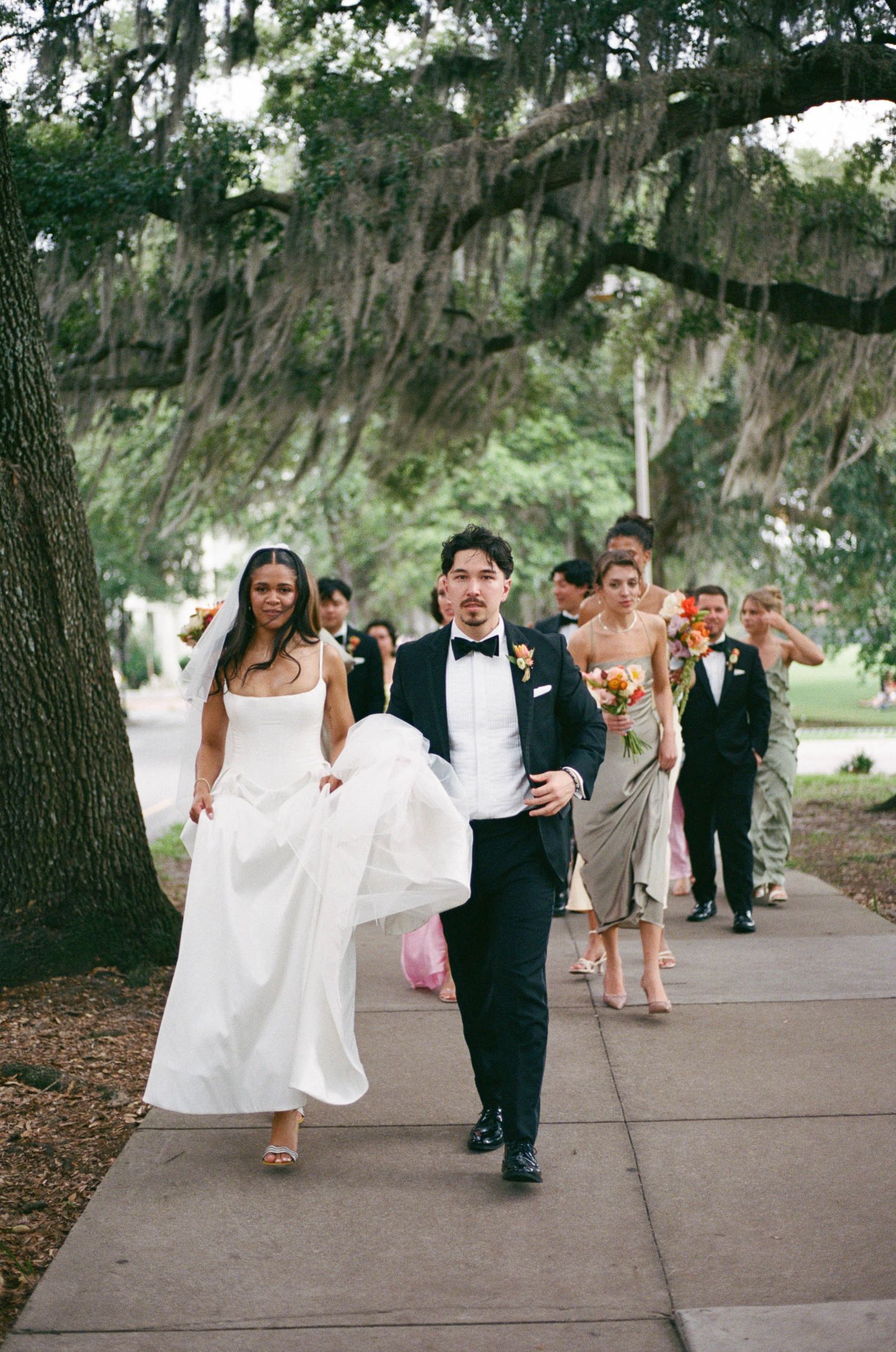Wedding party walking along a tree-lined path with bridesmaids in pastel dresses and white flowers.