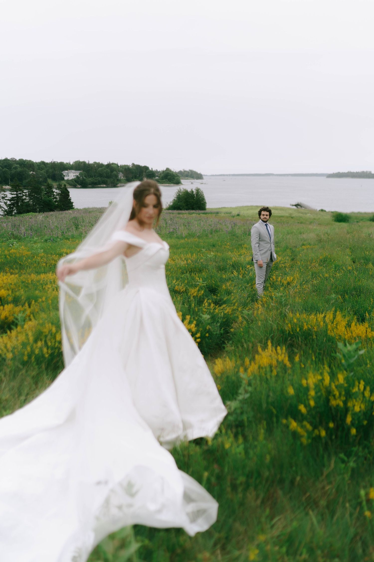 A wedding couple poses in a meadow of yellow flowers near a waterfront on an overcast day.