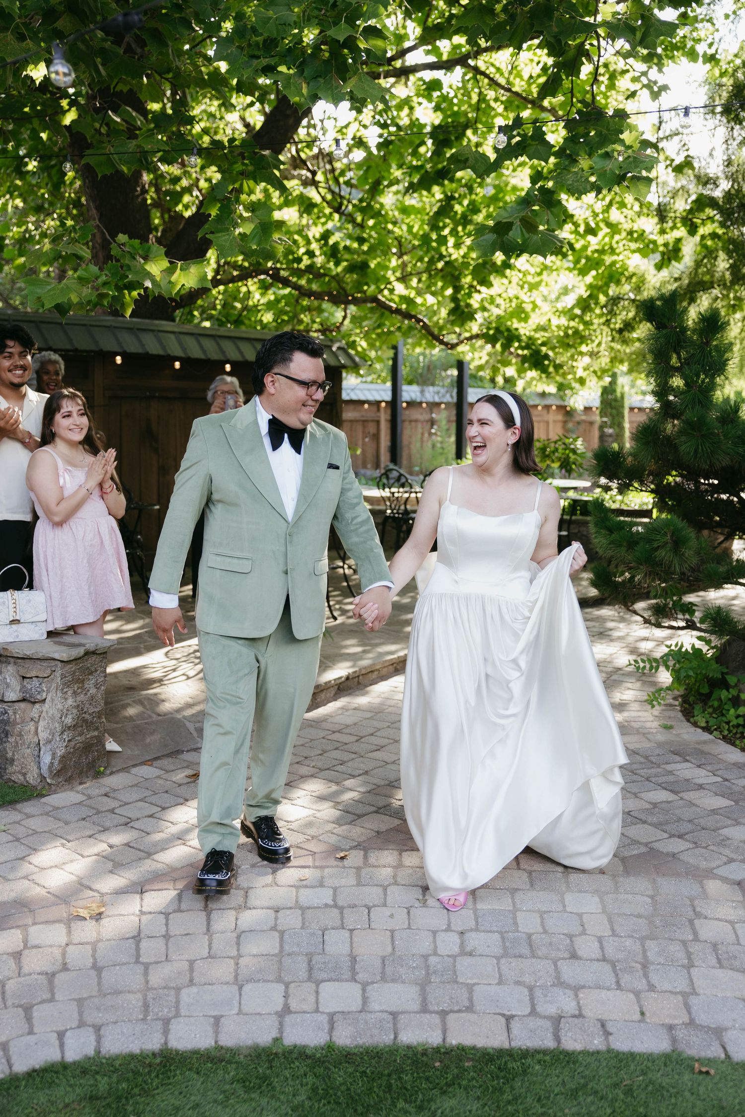 A couple in wedding attire dances and twirls on a stone patio surrounded by greenery and trees.