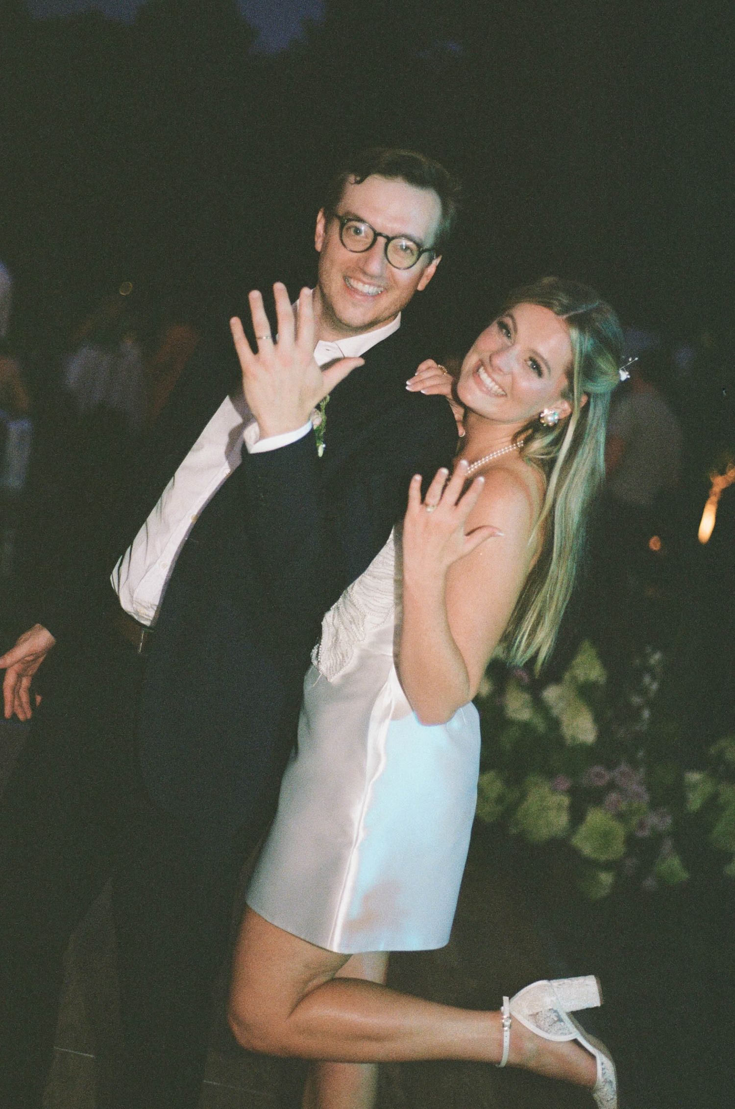 Newlywed couple smiling and showing wedding rings at outdoor evening reception, bride in white mini dress.