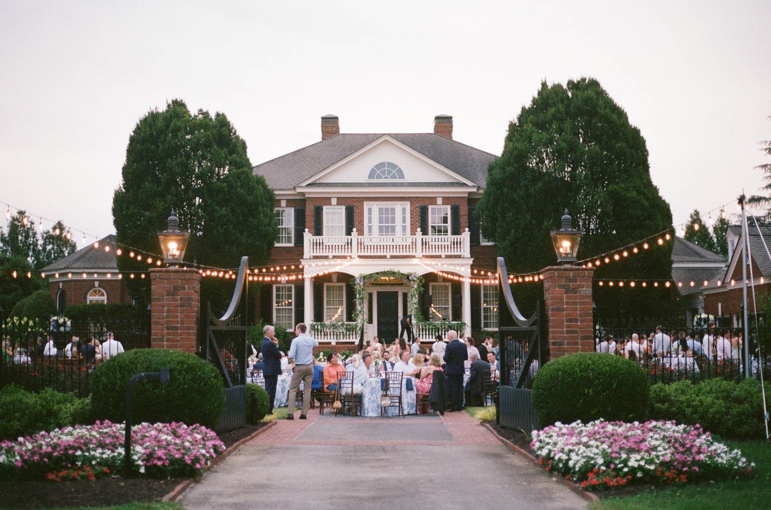 Elegant Southern mansion wedding venue with string lights, lush greenery, and guests gathered at dusk for an outdoor ceremony.