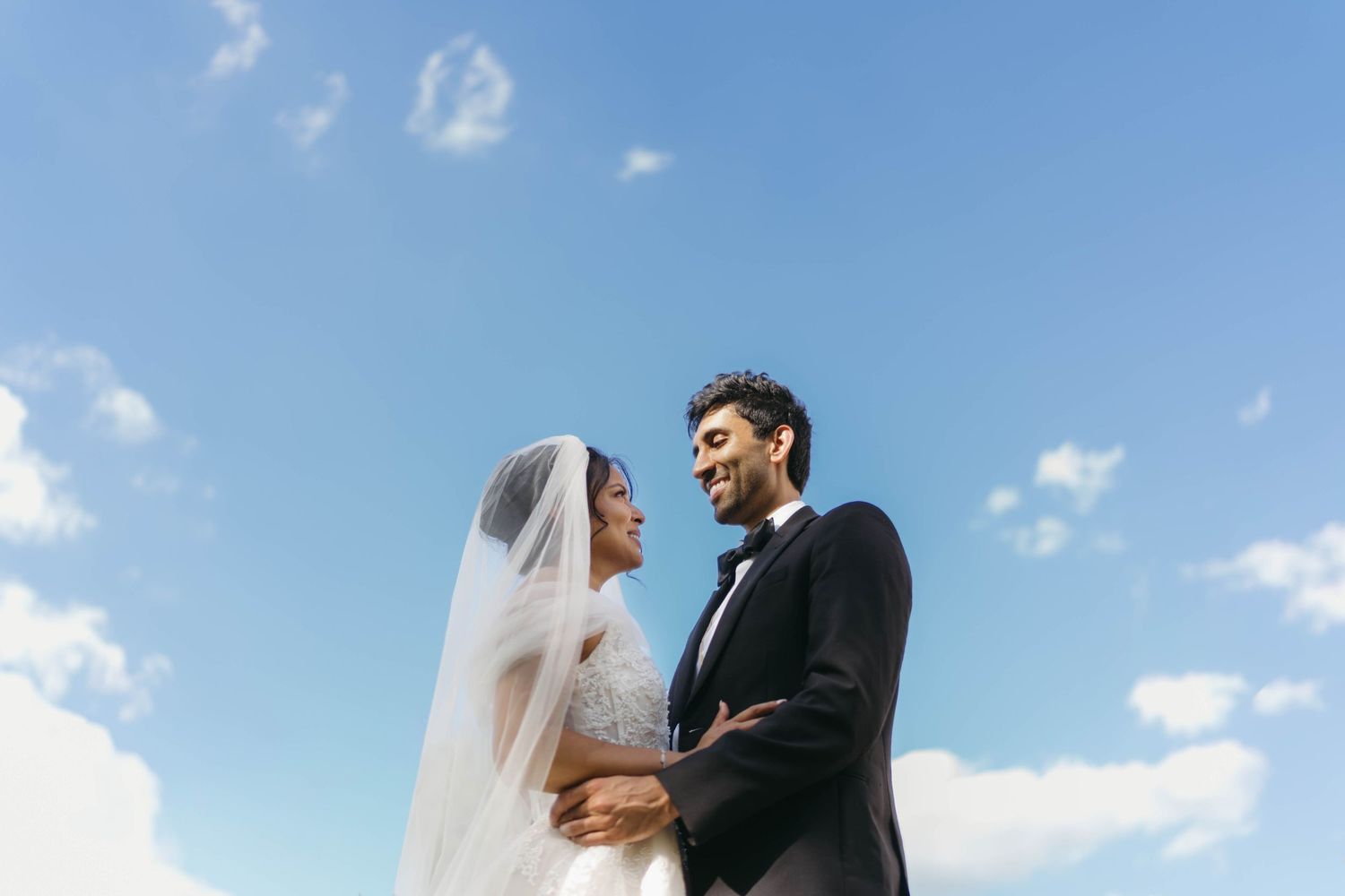 Bride and groom share romantic moment under blue sky, bride wearing veil and groom in tuxedo.