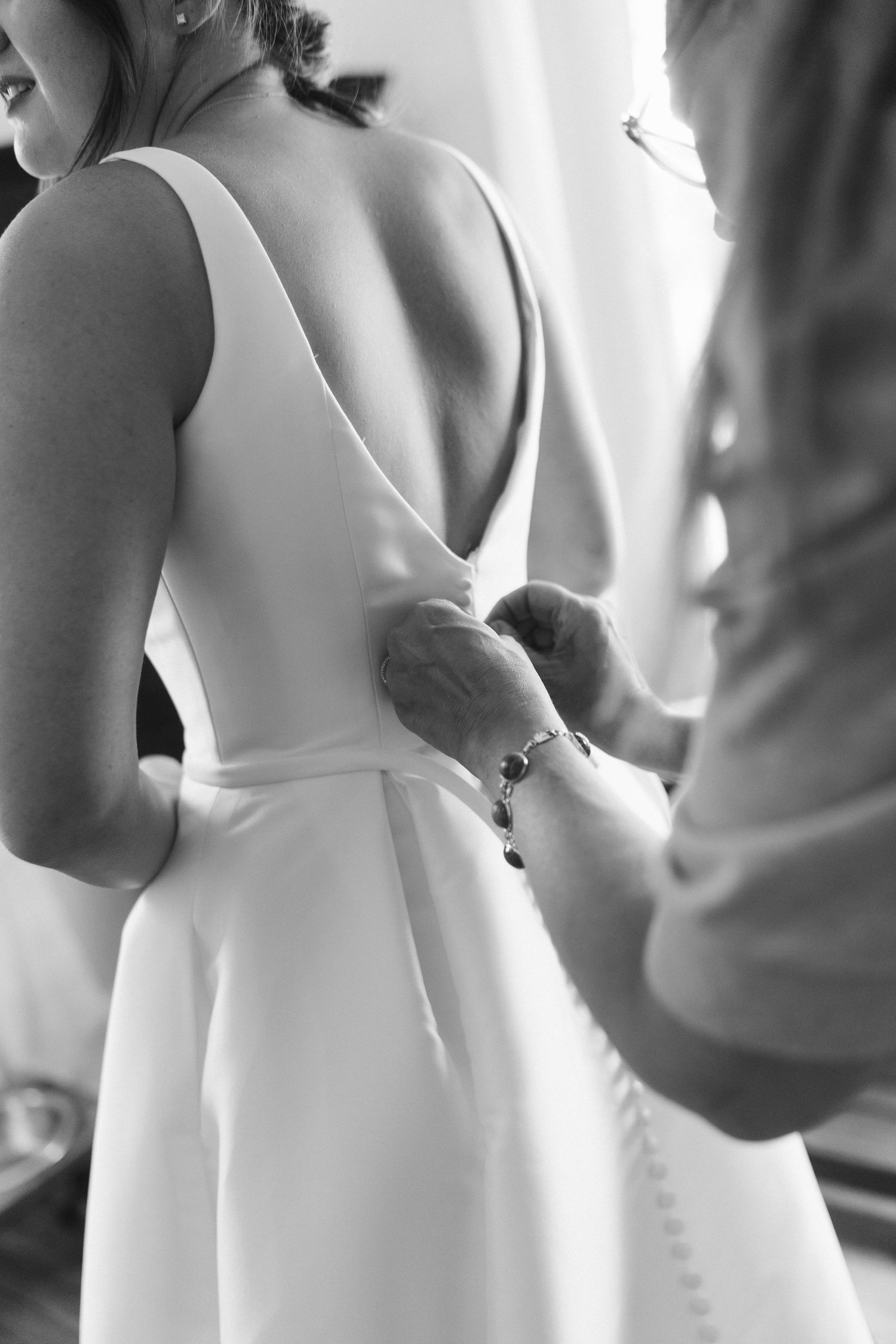 Close-up black and white photo of hands fastening buttons on the back of an elegant white wedding dress.