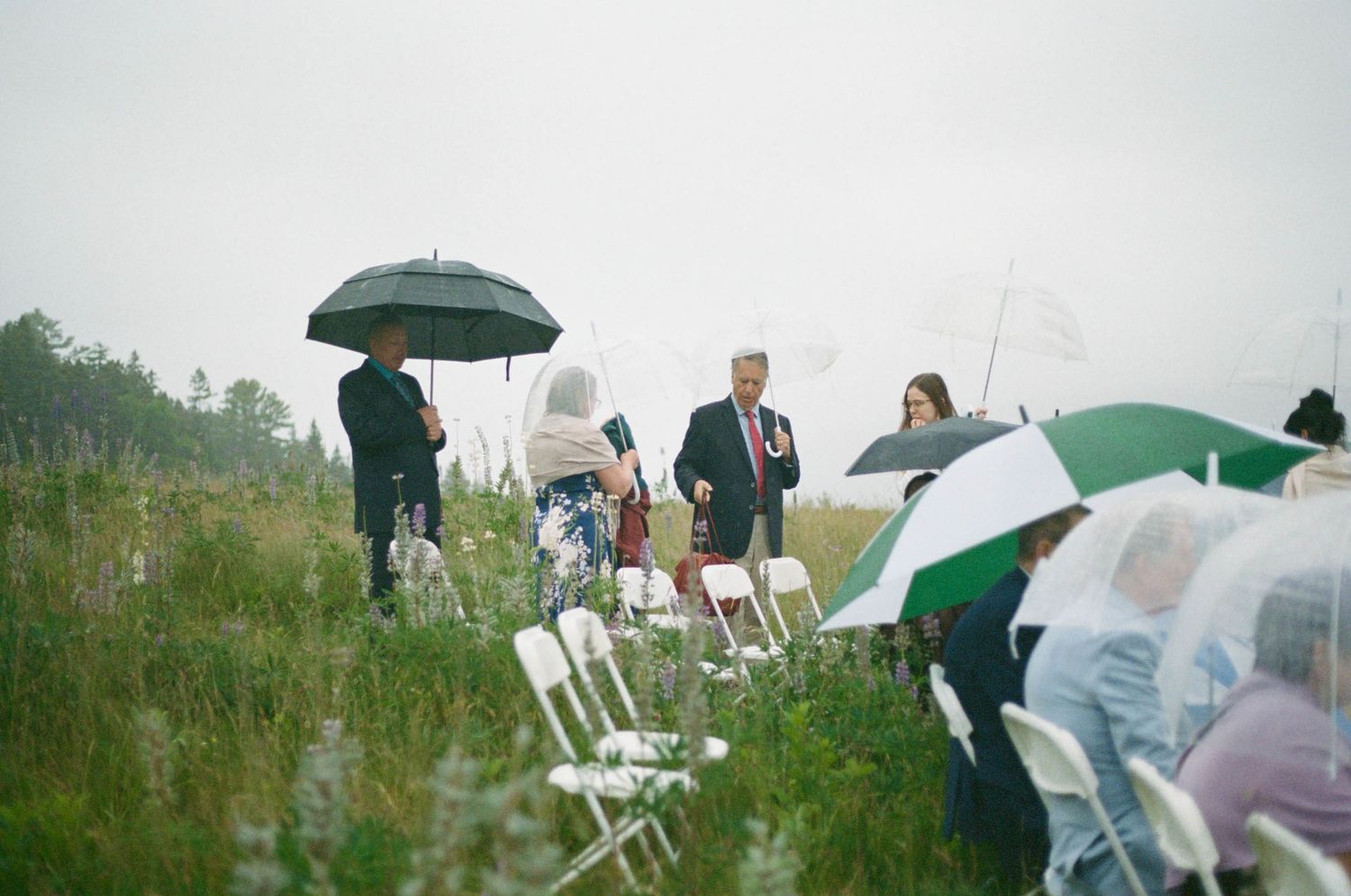 Wedding guests huddle under green umbrellas in a misty field with white folding chairs.