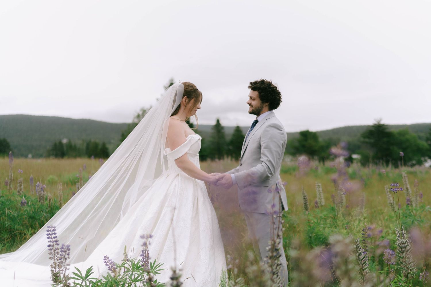 A couple walks through a misty field of purple wildflowers holding a white umbrella.