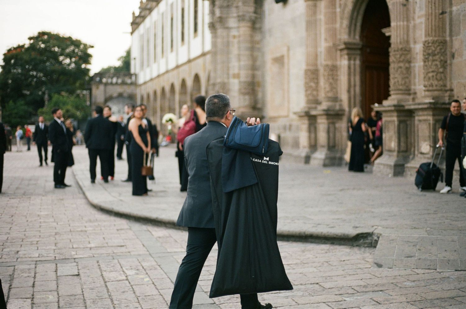 People in formal attire gathering outside a historic colonial building with stone archways.