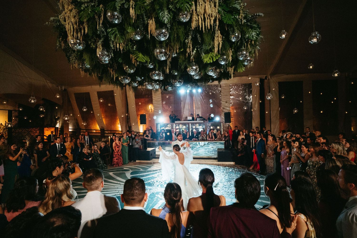 Wedding guests gather around a dance floor at an evening reception.