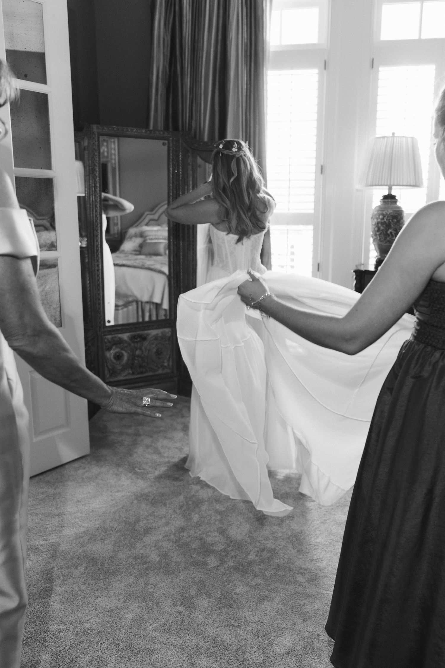 Black and white photo of bride being helped into white wedding gown in front of mirror.