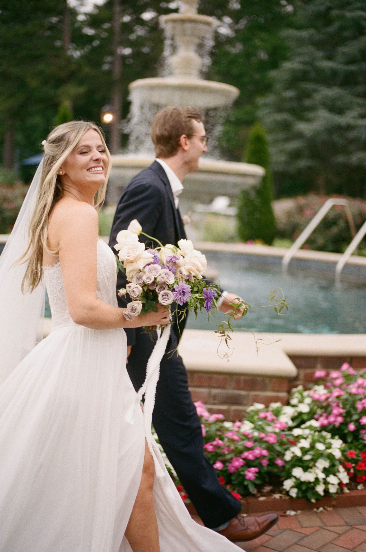 Smiling bride in white gown holding purple floral bouquet walks with groom near stone fountain.