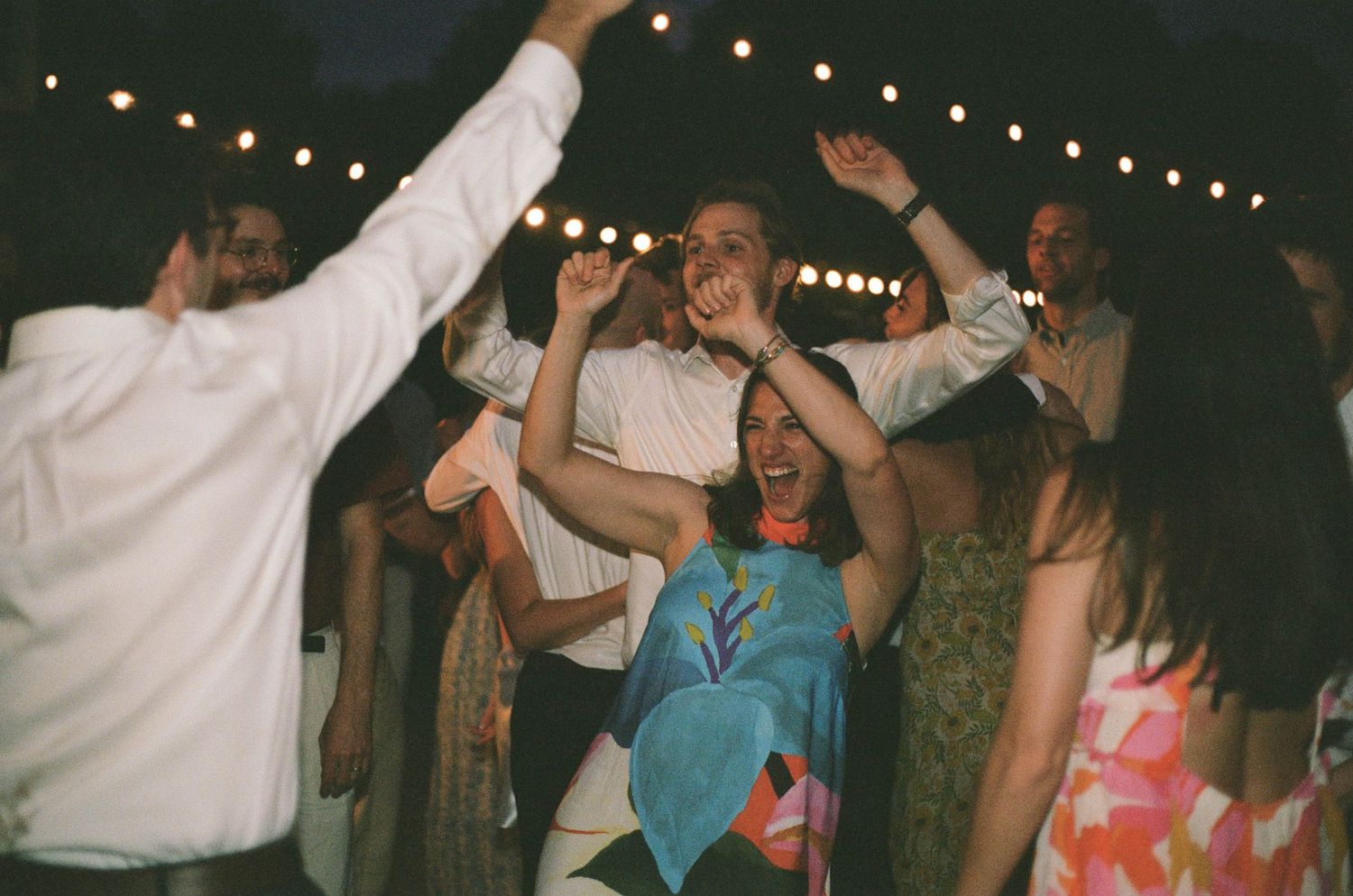 Wedding guests dance joyfully under string lights at an outdoor evening reception party.