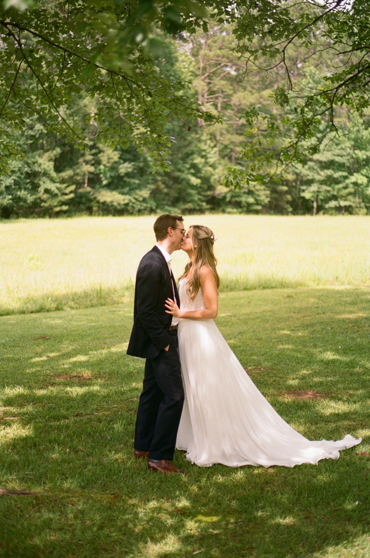 Bride and groom share romantic kiss on lush green lawn surrounded by trees.