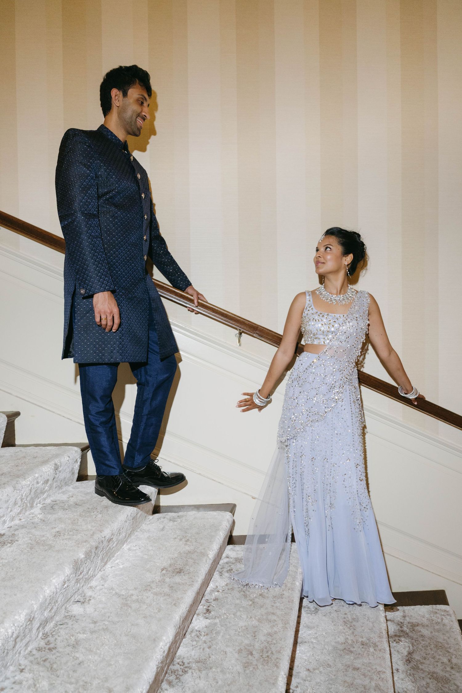 Couple in Indian wedding attire on marble staircase, man in navy sherwani, woman in silver embellished saree.