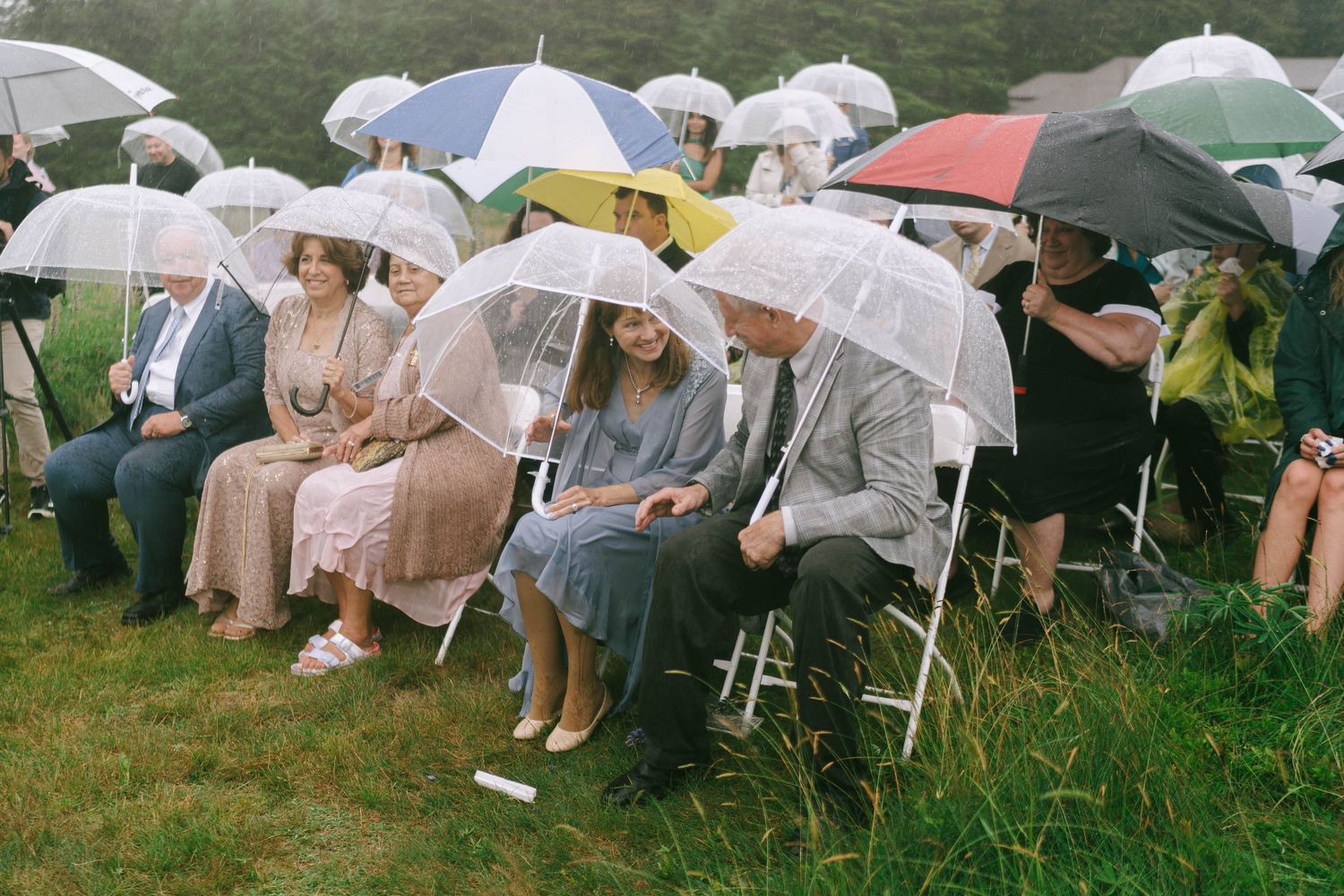 Wedding guests hold white umbrellas while seated on folding chairs outdoors.