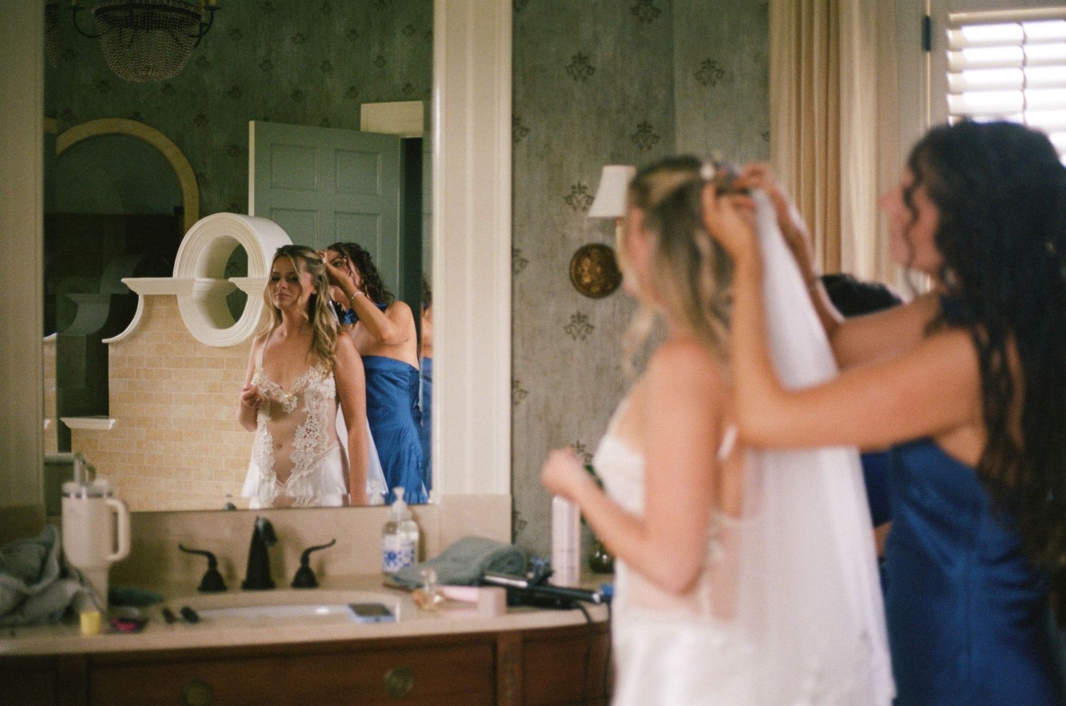 Bridesmaids helping bride with hair and veil in vintage mirror dressing room before wedding ceremony.