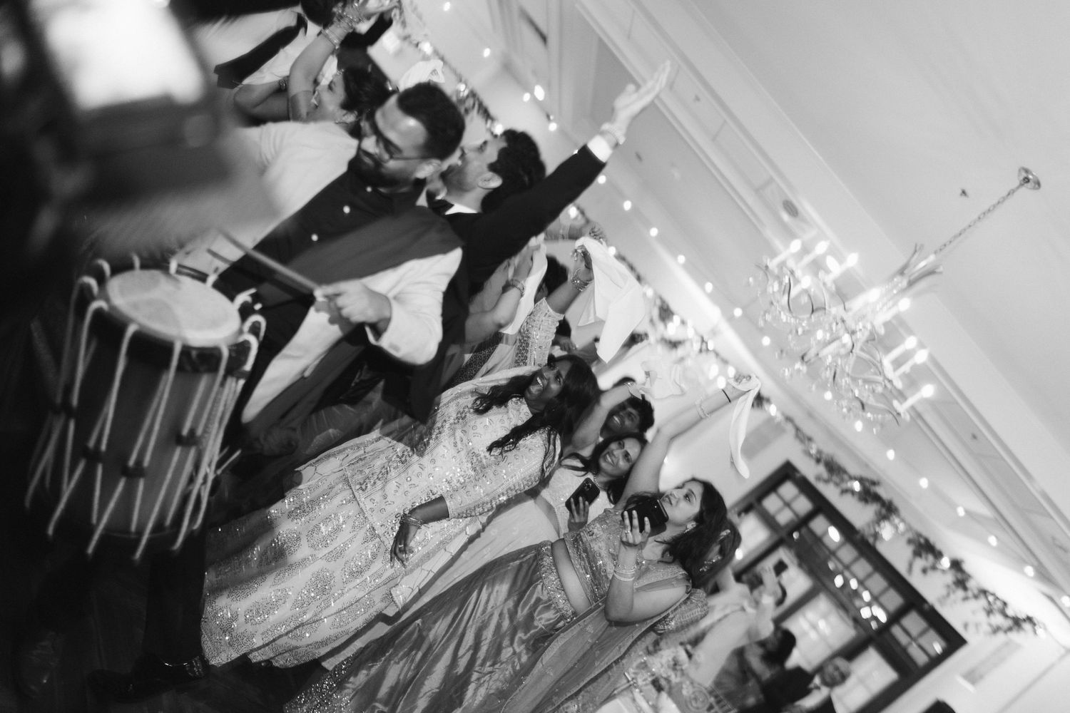 Black and white photo of wedding guests dancing with dhol drummer at Indian reception.