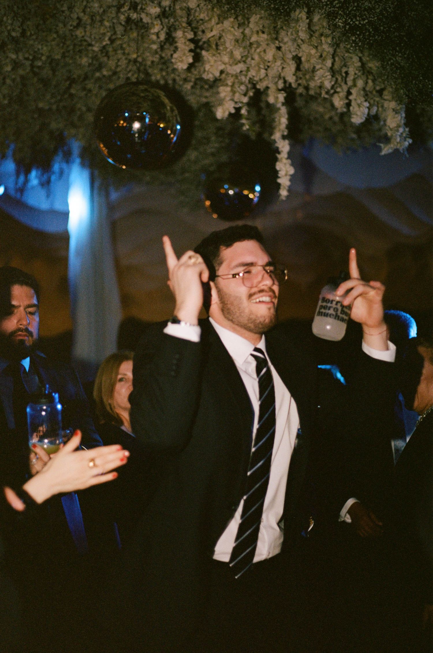 Wedding reception guests dancing and celebrating under decorative floral ceiling arrangements with blue lighting.