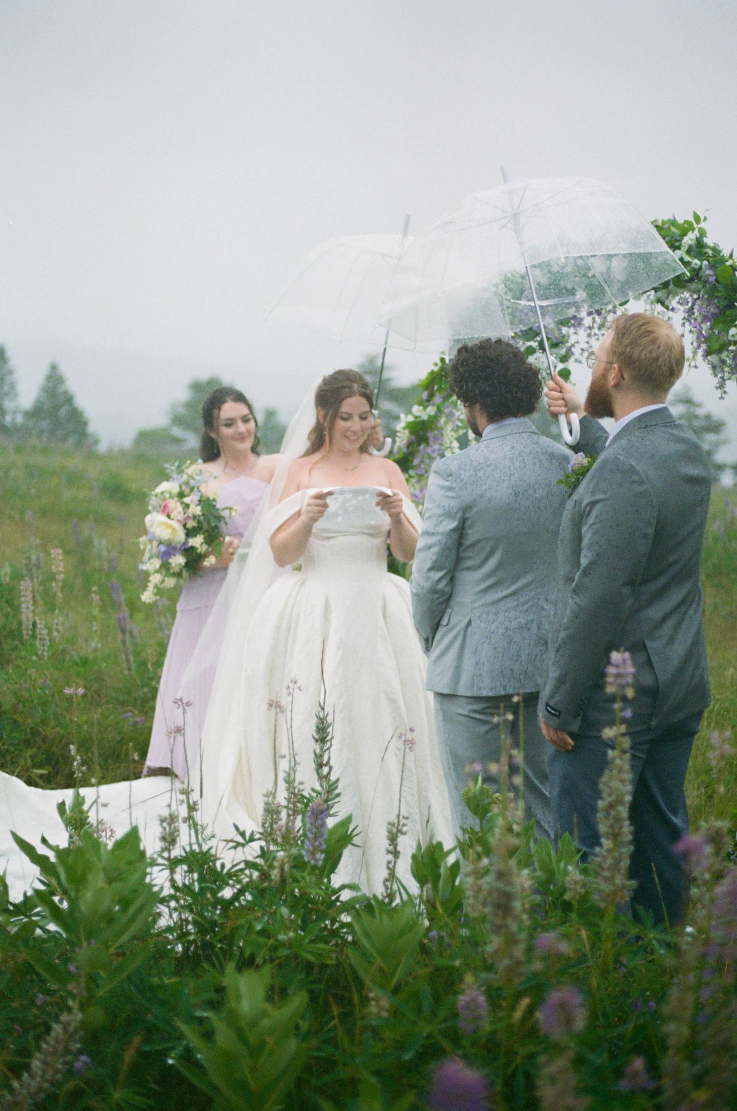 Intimate wedding ceremony surrounded by wildflowers and clear umbrellas on overcast day.