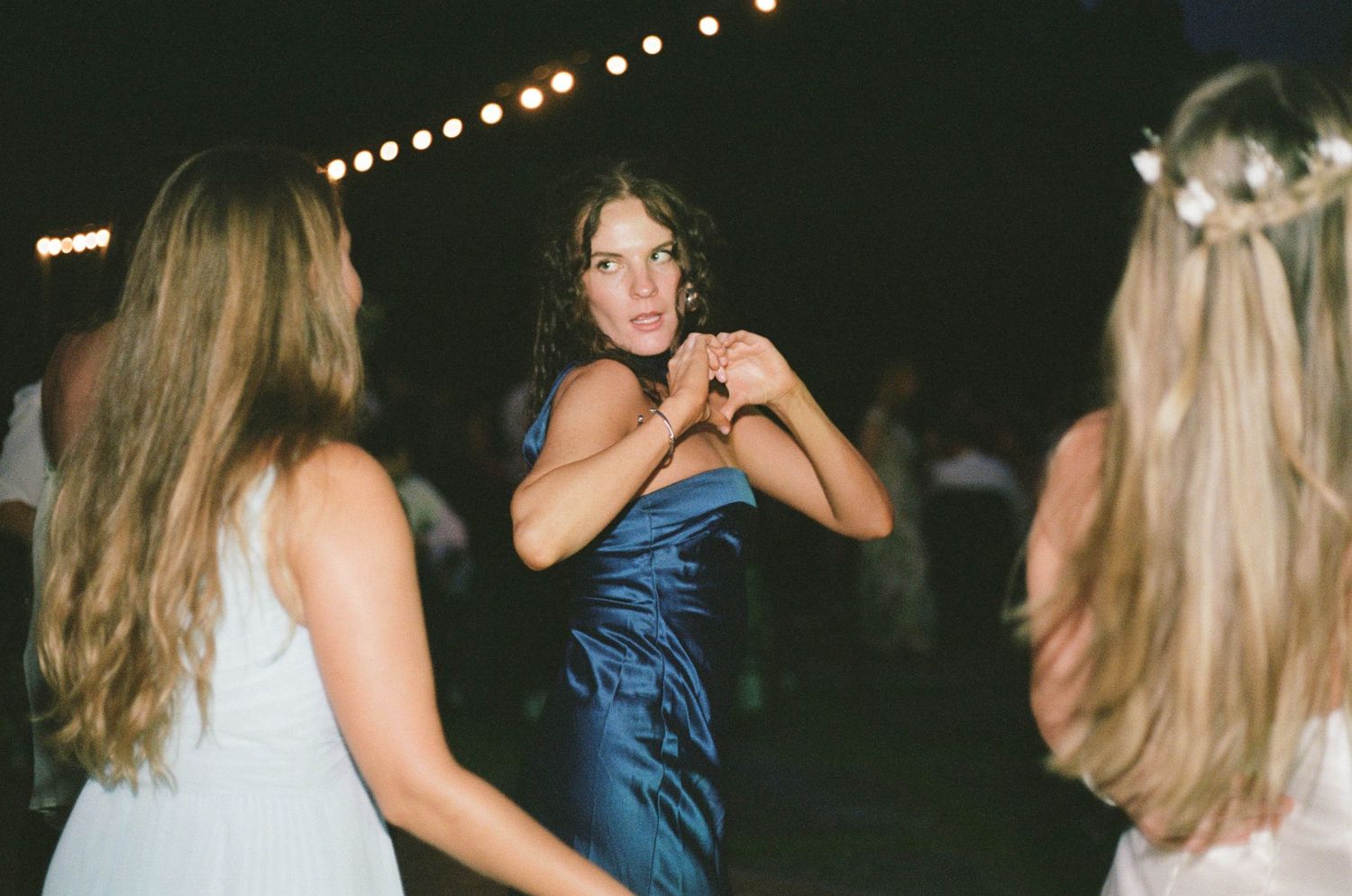 Woman in blue satin dress dances energetically at an outdoor wedding reception under string lights.