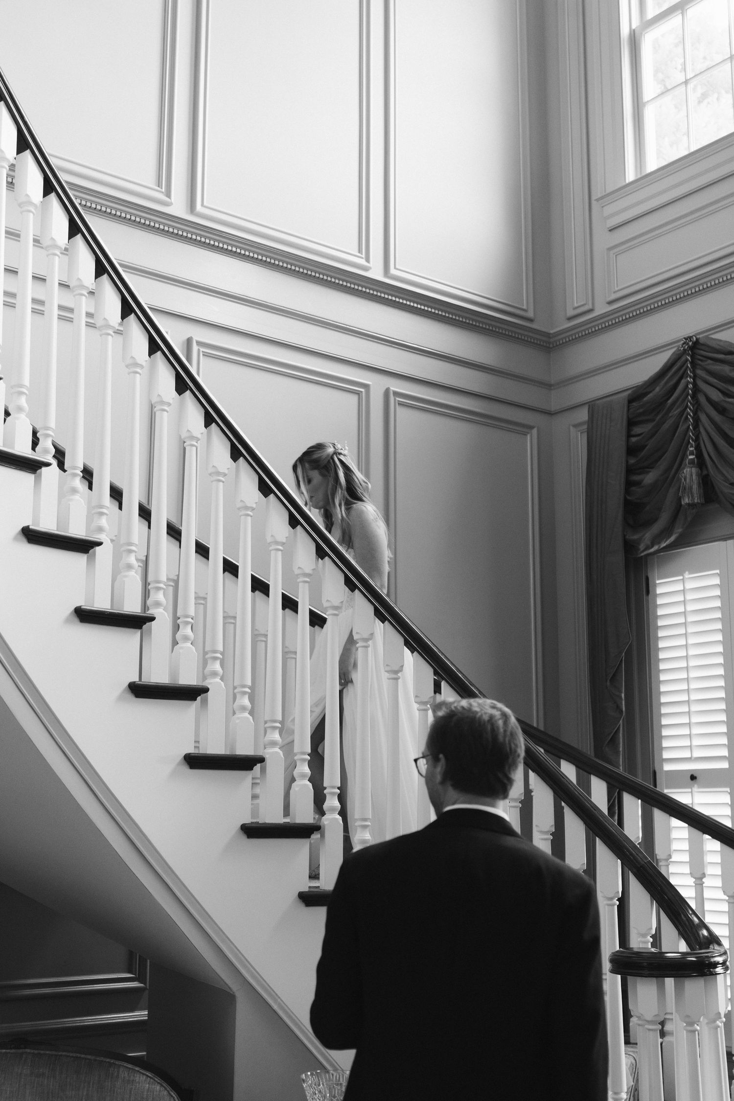 Black and white photo of bride descending elegant staircase as groom waits below.