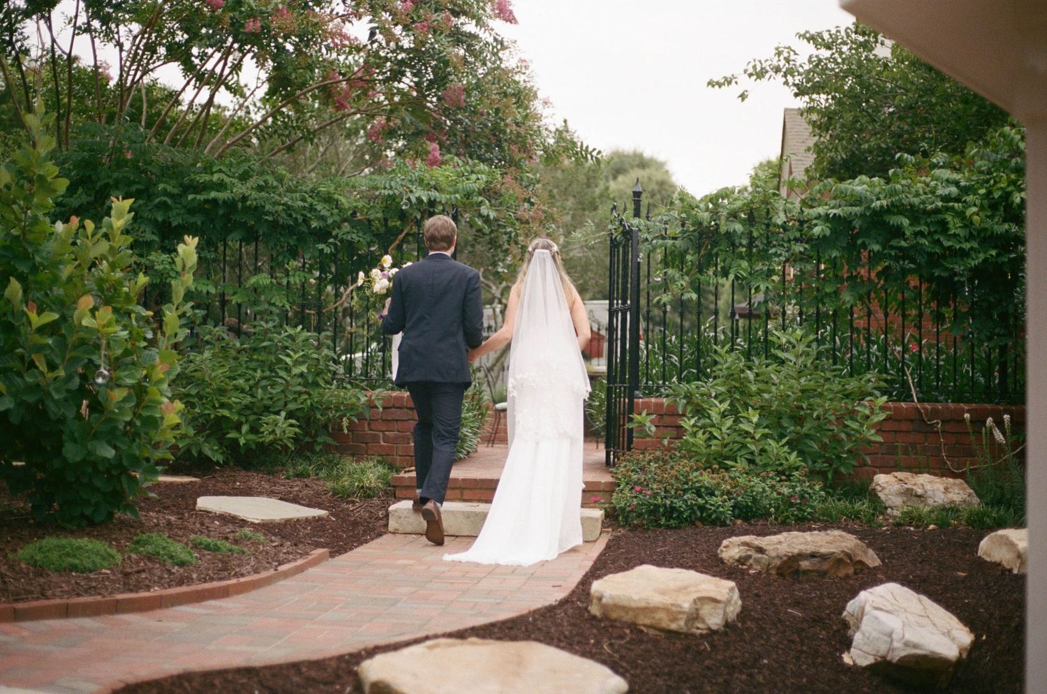 Bride and groom walking hand in hand through a lush garden pathway toward an iron gate on their wedding day.