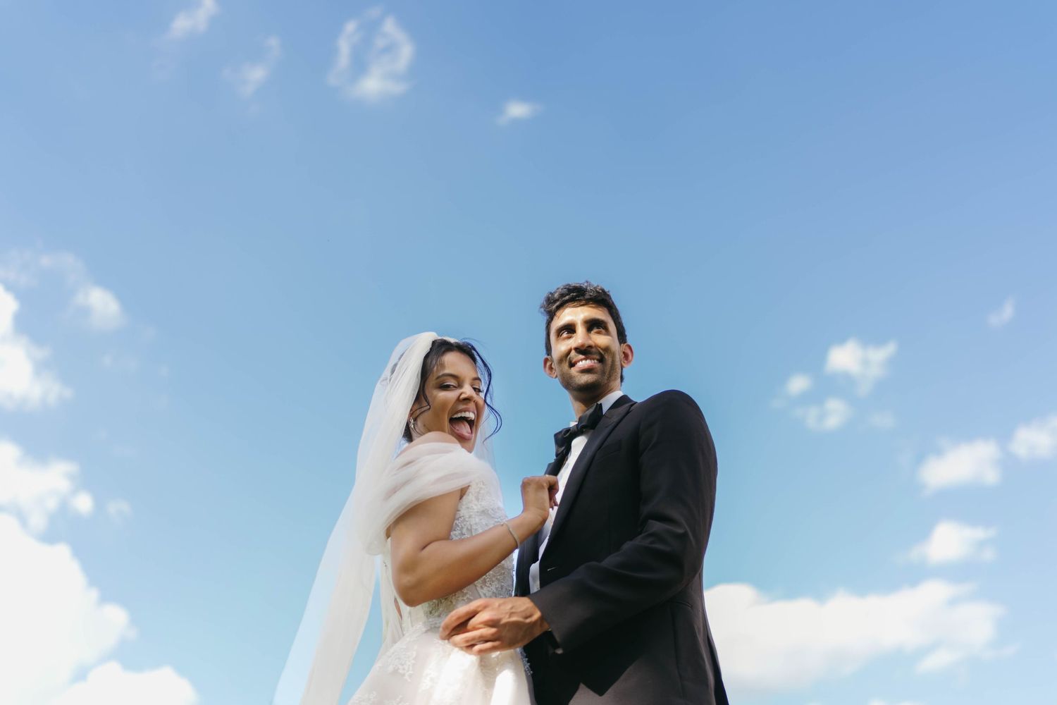 Laughing bride and groom embrace under bright blue sky with white clouds on their wedding day.